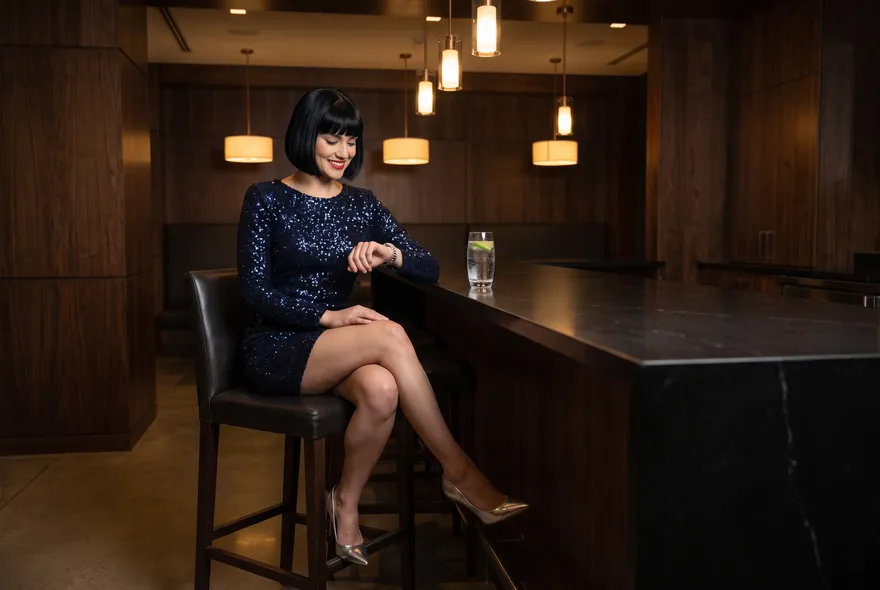 The same woman sits on a tall stool at a dark stone counter in a dim lounge, smiling at her silver wristwatch, a glass of sparkling water beside her.